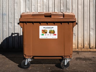 Brūna atkritumu tvertne uz riteņiem ar uzlīmi, kas vēsta "BIO ATKRITUMI" un "VEĢETĀRIE UN DĀRZA ATKRITUMI" ar attēliem par to, ko drīkst un ko nedrīkst tajā mest. Tvertne novietota uz asfalta pie gofrētas metāla sienas.