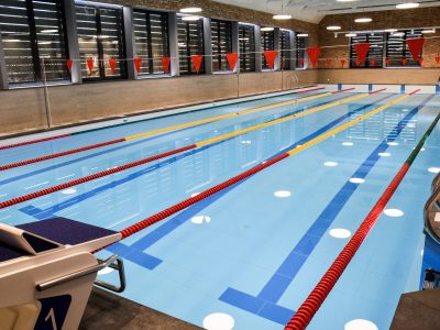 Kuldīgas peldbaseins. Iekštelpu peldbaseins ar gaiši zilu ūdeni, vairākām zilām celiņu līnijām un sarkani dzeltenām celiņu virvēm. Priekšplānā redzams starta bloks ar numuru "1", bet virs baseina izkārtoti sarkani karodziņi.