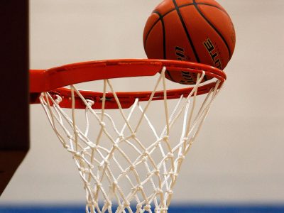 Tuvplānā redzama oranža basketbola bumba, kas atrodas uz sarkanas groza stīpas ar baltu tīklu. Fonā gaiša siena ar zilu svītru.