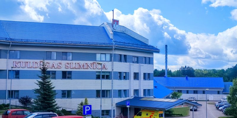 Kuldīgas slimnīcas galvenā ēka ar ziliem jumtiem un gaišām fasādēm. Uz ēkas ir uzraksts "KULDĪGAS SLIMNĪCA".