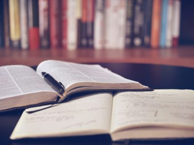 Atvērta grāmata ar melnu pildspalvu un atvērta piezīmju grāmatiņa ar rokrakstu uz galda, fonā aizmigloti grāmatu plaukti.