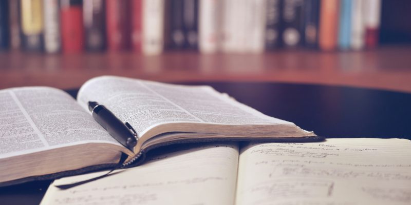 Atvērta grāmata ar melnu pildspalvu un atvērta piezīmju grāmatiņa ar rokrakstu uz galda, fonā aizmigloti grāmatu plaukti.