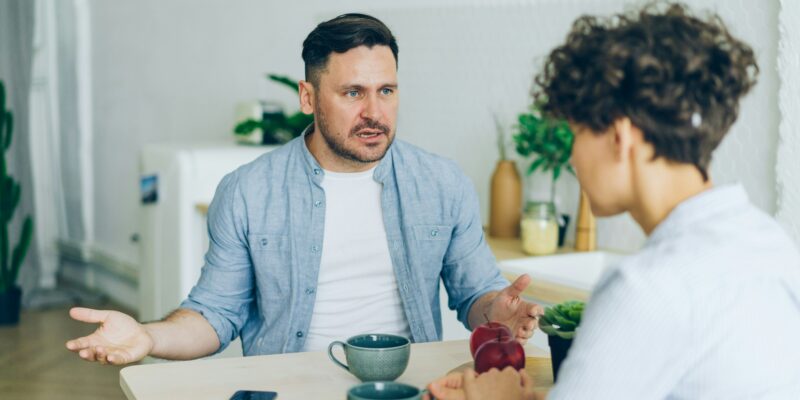 Vīrietis gaiši zilā kreklā, sēžot pie galda, runā ar sievieti, kas sēž viņam pretī, un izteiksmīgi žestikulē ar rokām. Uz gaišā koka galda ir krūzes, sarkani āboli un viedtālrunis.