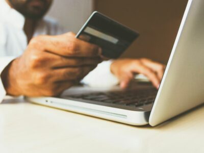 Cilvēks ar tumšu bankas karti vienā rokā un pirkstiem uz klēpjdatora tastatūras, visticamāk, veic tiešsaistes maksājumu.