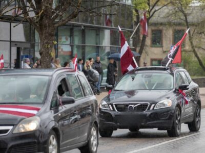 Mitrā ielā brauc divas melnas automašīnas, rotātas ar Latvijas karogiem. Priekšējais melnais apvidus auto uz jumta nes divus Latvijas karogus un sarkanmelnu karogu ar simbolu, uz tā sāniem ir uzraksts "Paldies visiem". Aizmugurējā automašīna ir daļēji redzama ar sarkanbaltsarkanu lentu. Fonā, uz ietves, stāv cilvēku pūlis, daži zem lietussargiem, vērojot notiekošo lietainā laikā.
