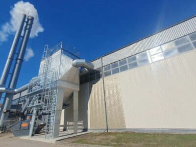 Industriāla ēka ar diviem augstiem pelēkiem skursteņiem, no viena nāk dūmi. Pie ēkas redzama ventilācijas sistēma un pelēks atkritumu konteiners ar oranžu uzrakstu "ALKO" gaiši zilās debesīs ar vienu mākoni.