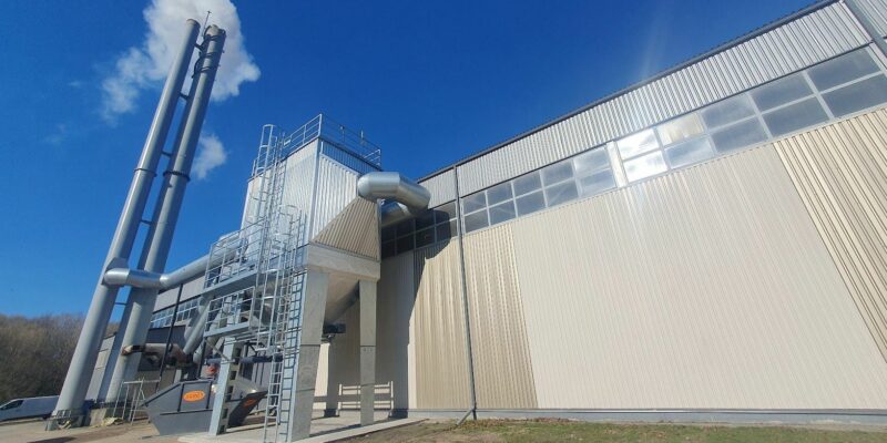 Industriāla ēka ar diviem augstiem pelēkiem skursteņiem, no viena nāk dūmi. Pie ēkas redzama ventilācijas sistēma un pelēks atkritumu konteiners ar oranžu uzrakstu "ALKO" gaiši zilās debesīs ar vienu mākoni.