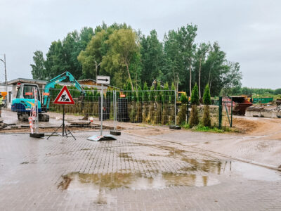 Lietainā dienā slapja bruģēta teritorija ar peļķēm. Aiz pagaidu metāla žoga strādnieki dzeltenās vestēs pie zila mini-ekskavatora ar uzrakstu "RŪTA" un asfalta kaudzēm. Blakus stāv ceļa zīme "Darbi uz ceļa" (sarkans trīsstūris ar strādnieka simbolu) un balta bultas zīme, kas norāda pa labi. Fonā redzamas koku audzes, industriālas ēkas un lieli būvgružu konteineri.