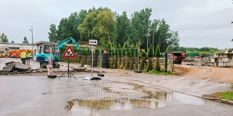 Lietainā dienā slapja bruģēta teritorija ar peļķēm. Aiz pagaidu metāla žoga strādnieki dzeltenās vestēs pie zila mini-ekskavatora ar uzrakstu "RŪTA" un asfalta kaudzēm. Blakus stāv ceļa zīme "Darbi uz ceļa" (sarkans trīsstūris ar strādnieka simbolu) un balta bultas zīme, kas norāda pa labi. Fonā redzamas koku audzes, industriālas ēkas un lieli būvgružu konteineri.