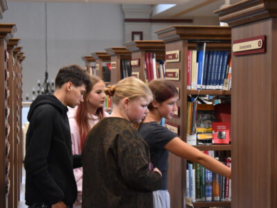 Četri jaunieši, divas meitenes un divi zēni, aplūko grāmatas koka grāmatu plauktos bibliotēkā. Meitene labajā pusē sniedzas pēc grāmatas, kas atrodas zemākā plauktā. Uz viena no plauktiem ir uzraksts "Literatūrmīlne".