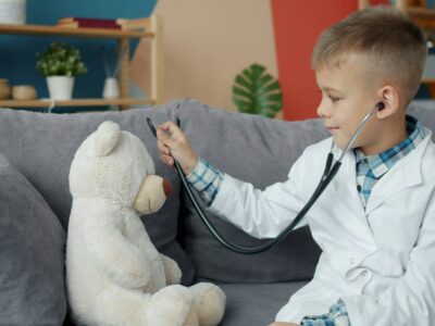 Zēns baltā ārsta halātā ar stetoskopu klausās plīša lācīša krūtis, spēlējoties dakteros uz dīvāna.