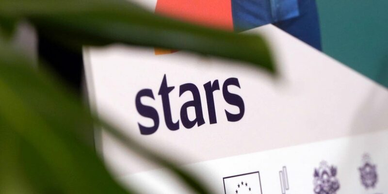 Balts plakāts vai stends ar vārdu "stars" tumši violetā krāsā. Zem tā redzams ES karoga logo, skaitlis "2027" un divi ģerbonim līdzīgi logo. Daļēji aizsegts ar zaļu augu lapām.