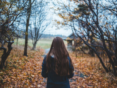 Persona no mugurpuses ar gariem brūniem matiem iet pa rudens taku, kas noklāta ar kritušām oranžbrūnām lapām, ar kailiem kokiem un lauku koka būdiņu fonā.