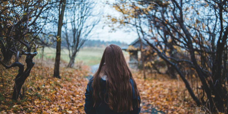 Persona no mugurpuses ar gariem brūniem matiem iet pa rudens taku, kas noklāta ar kritušām oranžbrūnām lapām, ar kailiem kokiem un lauku koka būdiņu fonā.